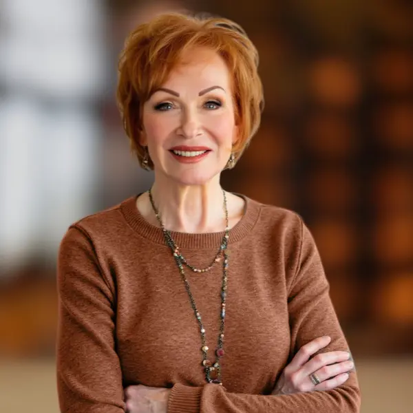 A confident, mature woman with short red hair smiling with arms crossed, wearing a brown sweater and necklace, standing in a softly blurred indoor setting.